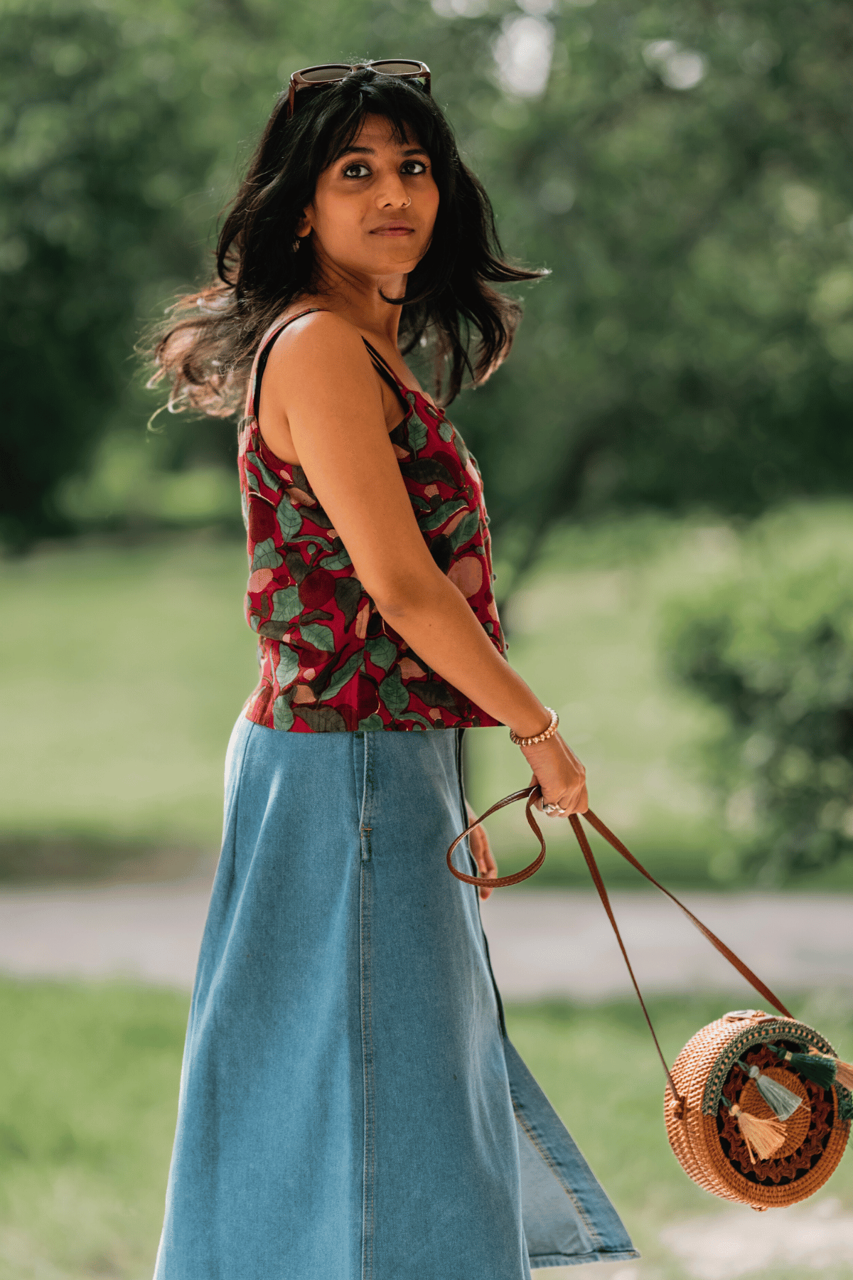 Woman in a floral top and denim skirt holding a round woven bag outdoors.